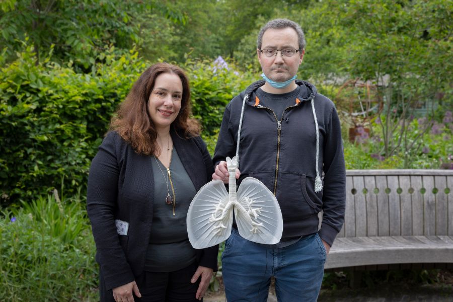 Two people standing in a garden smiling at the camera. The person on the left has long brown hair, is wearing a black blazer/cardigan and is wearing a dark green shirt. The person on the right is wearing glasses and a face mask pulled down from his mouth, is wearing a black zipped jacket and dark blue jeans. The person on the right is holding an origami replica of a pair of lungs.